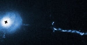 A deep space image of a bright galaxy with a luminous blue core on the left, extending a faint, irregular jet of light to the right. The background is a dark, star-speckled sky.