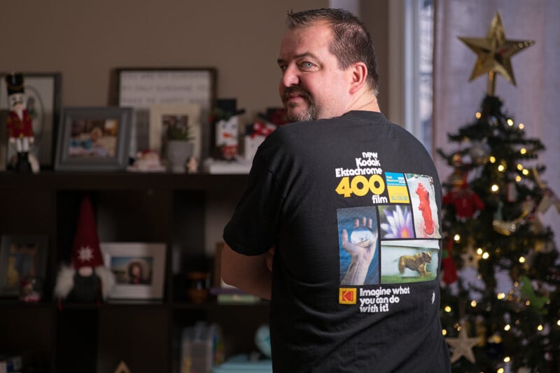 A man faces slightly away, wearing a Kodak Ektachrome 400 film T-shirt. Behind him is a decorated Christmas tree with lights and ornaments. Shelves are filled with various items, including a nutcracker and framed photos.