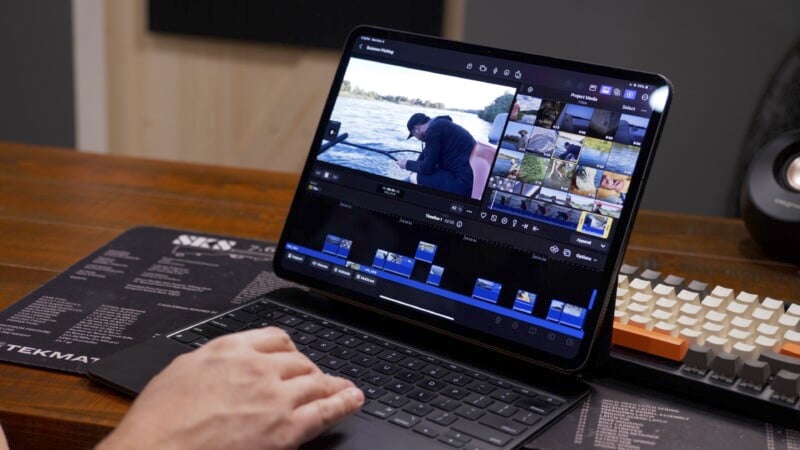 A person using a tablet with a keyboard on a wooden desk, editing a video. The video editing software displays clips of a man near water. A mechanical keyboard and a speaker are in the background.