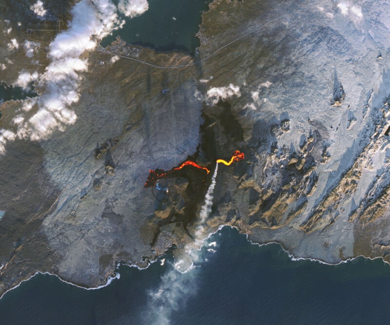 Aerial view of a volcanic landscape with a bright, glowing lava flow cutting across dark terrain. Smoke rises from the lava, and nearby clouds hover above. The coastline is visible at the bottom of the image.