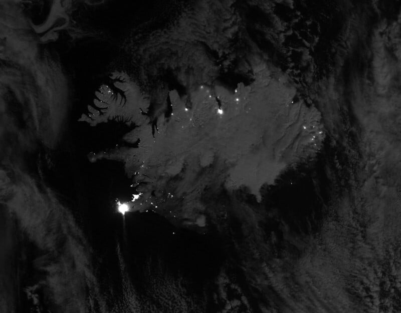 A satellite image of Iceland at night, showing the island's outline in grayscale. Bright city lights are visible, highlighting urban areas like Reykjavík. Surrounding clouds and water appear dark.