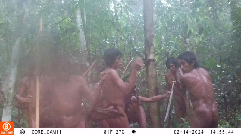 A group of people in a forest setting, holding tools or weapons, appear to be in conversation or engaged in an activity. The image is slightly blurry and shows a dense, green wooded area. The timestamp indicates the photo was taken in February 2024.