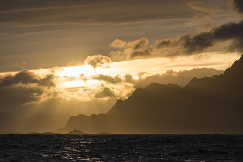 A dramatic sunset over a body of water with silhouetted mountain ​peaks. The sun breaks through thick clouds,casting golden ‍rays⁢ across the sky⁣ and reflecting on the waves below.