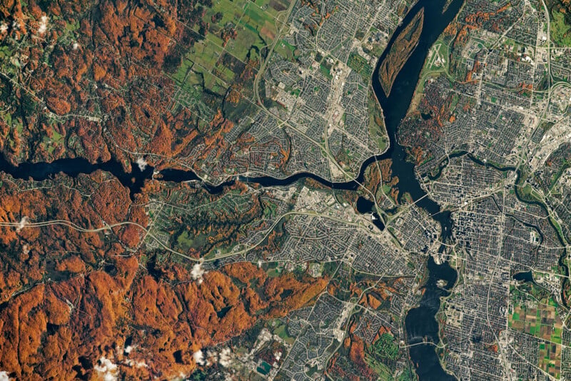 Aerial view of a city divided by a river. The left side shows dense red-orange autumn foliage, while the right side features urban development with roads and buildings. The river and bridges connect the contrasting landscapes.