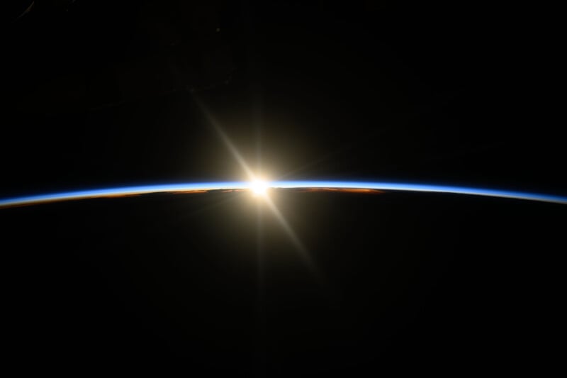 A stunning view of the sun rising over Earth's horizon, with a thin blue line of atmosphere visible against the blackness of space. The sun emits bright rays, creating a starburst effect.