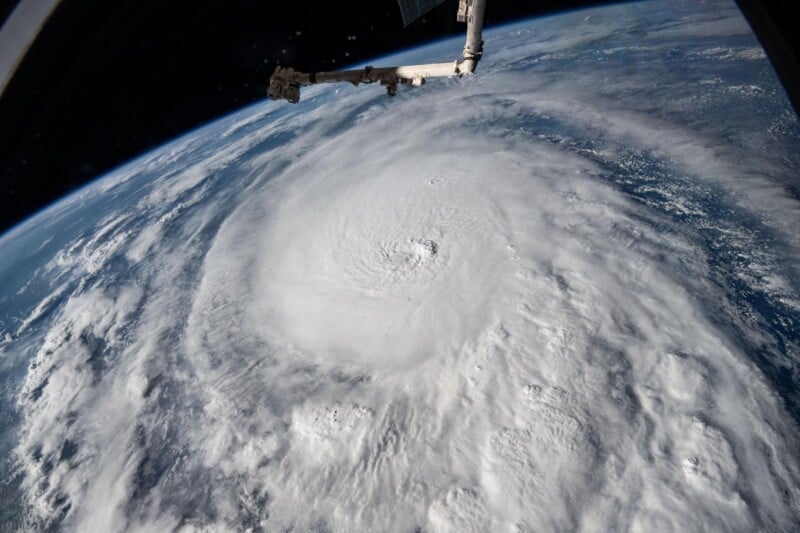 View of a massive hurricane from space, showing a swirling white cloud formation over the Earth. Part of a spacecraft is visible in the upper part of the image. The landscape below is partly obscured by clouds.