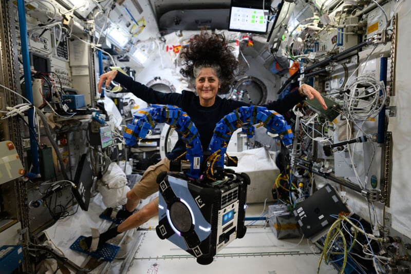 A person with long hair floating in a space station, wearing a black shirt and khaki shorts. They are interacting with a robotic device with blue appendages. The background is filled with equipment, wiring, and monitors in a high-tech environment.