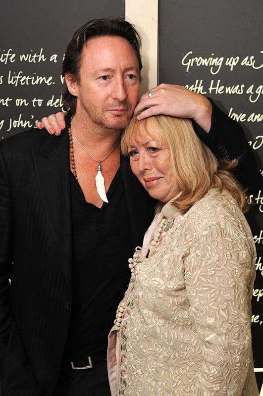 A man with dark hair and a necklace stands with his arm resting on the head of a woman with light hair. The woman wears a light-colored embroidered jacket. They pose in front of a black background with white cursive writing.