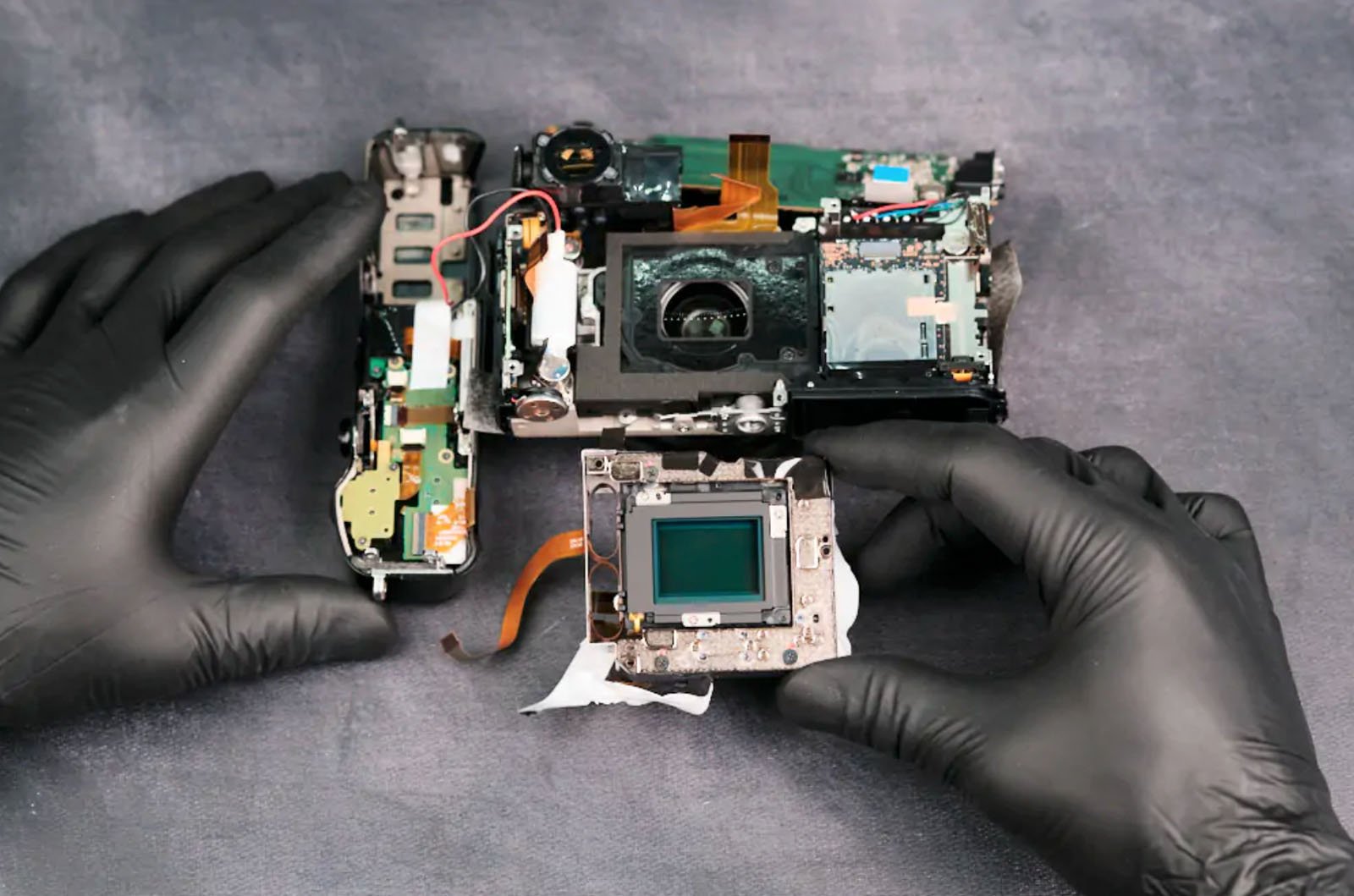 A person wearing black gloves disassembles a camera on a table. Various camera parts, including circuit boards and components, are spread out. The person is holding a camera sensor. The background is a textured, gray surface.