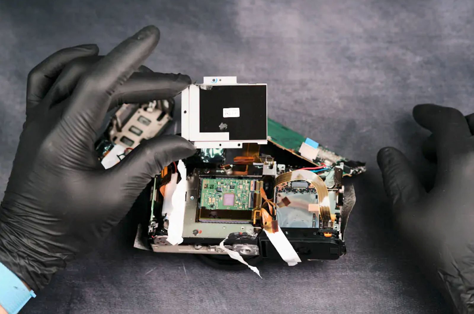 Hands wearing black gloves disassemble an electronic device, exposing its internal components like circuit boards and wiring. One hand holds a detached part above the device. The background is a textured gray surface.