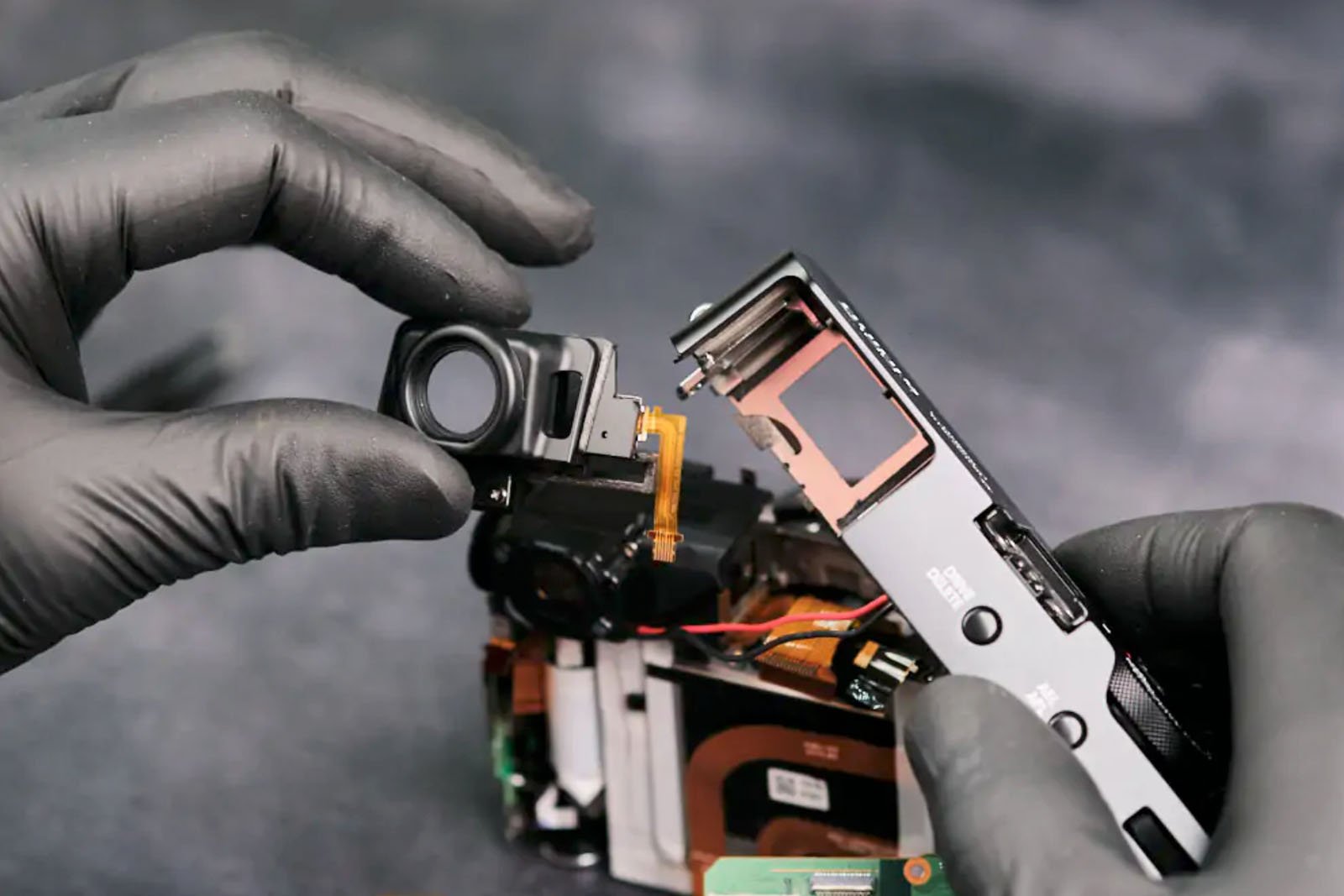 A person wearing black gloves disassembles a camera, holding a small electronic component apart from the camera body. The background is blurred, emphasizing the focus on the hands and camera parts.