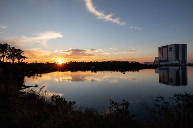 A tranquil sunset over a still lake reflects the vibrant hues of the sky. Silhouettes of trees frame the scene, and a large building with a NASA logo stands on the right, mirrored in the water.