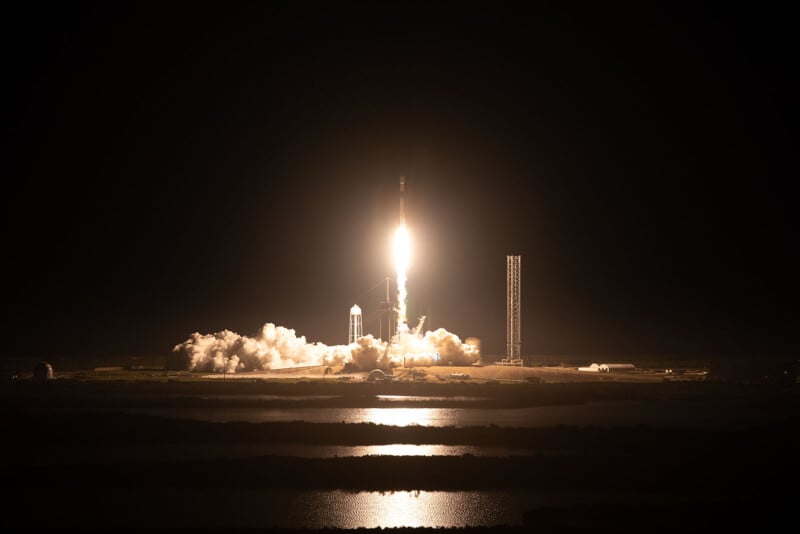 A rocket launches into the night sky, emitting bright flames and smoke. The dark surroundings highlight the luminous ascent, with a reflective body of water in the foreground.