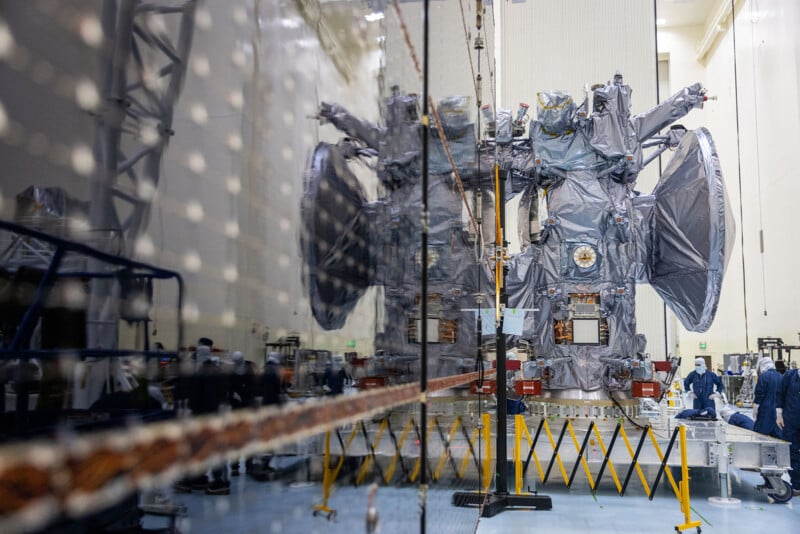 A spacecraft covered in metallic foil is in a clean room. It is reflected on a large, glossy surface. Several people in lab coats and hair nets work around it. The room is spacious and has industrial equipment and safety barriers.