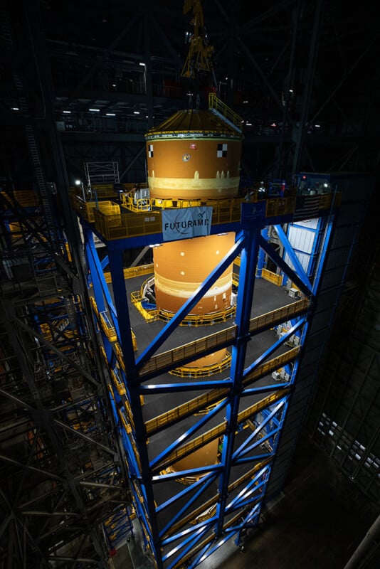 A large rocket booster stands vertically inside an industrial building. It's surrounded by blue support scaffolding, with platforms at various levels. The scene is dimly lit, emphasizing the booster and its size. A banner reads "Futurami.