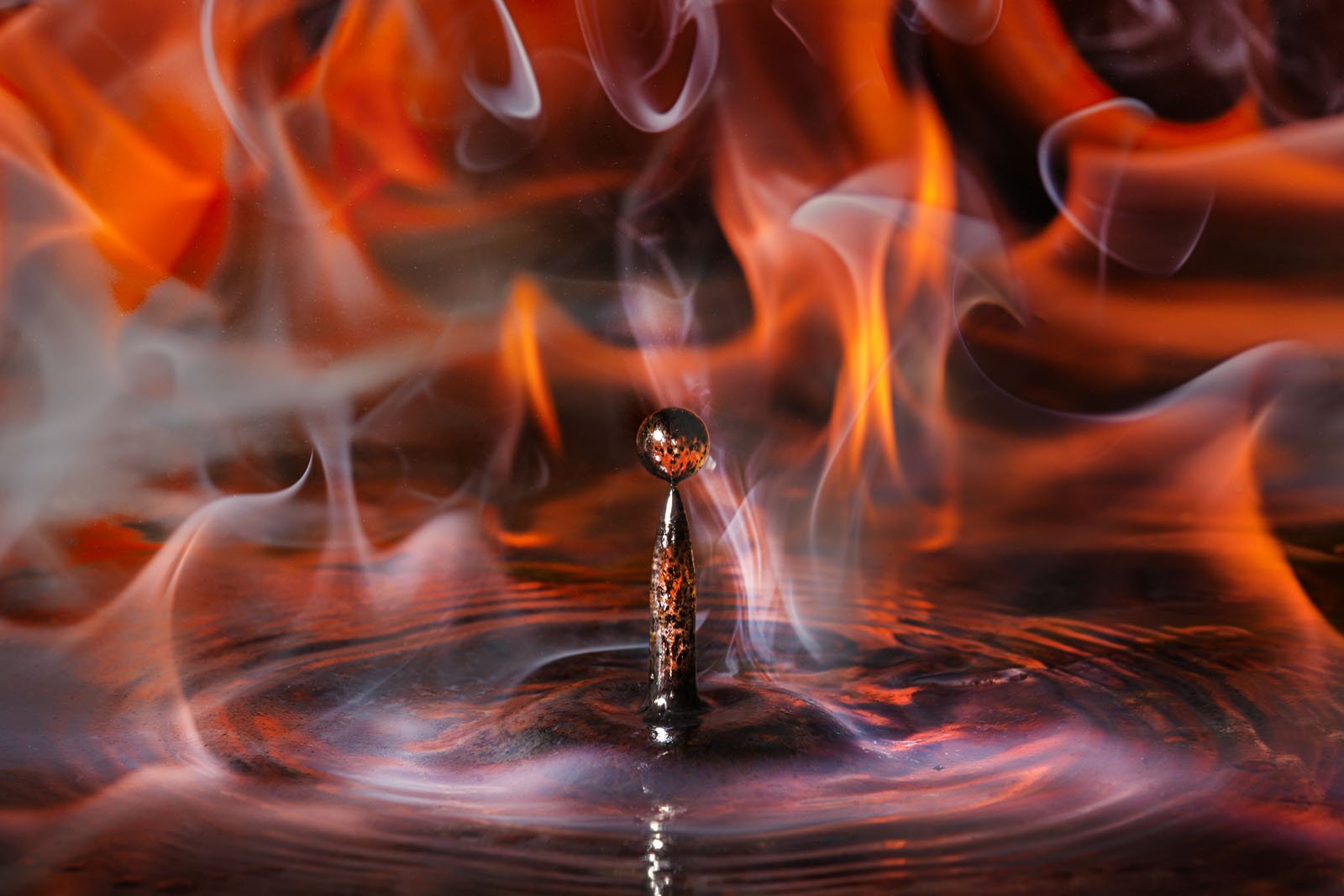 A water droplet rises above a surface, surrounded by swirling flames and smoke. The scene captures the contrast between the fluidity of water and the intense heat of fire, with ripples spreading outwards from the splash point.