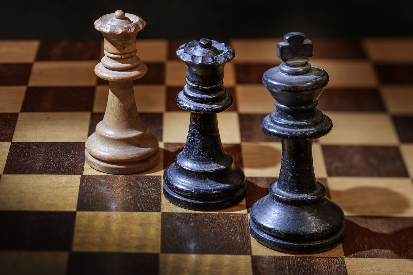 Close-up of a chessboard with three chess pieces: a wooden queen on the left and two dark-colored kings in the center and right. The board has alternating light and dark squares, with a dramatic lighting effect.