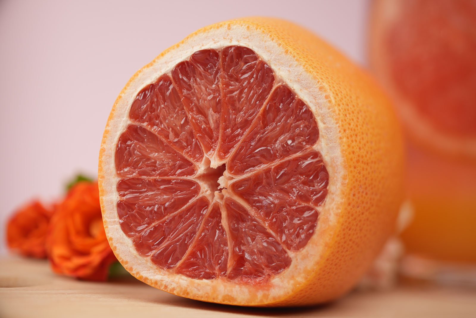 Close-up of a halved grapefruit with a vivid pink interior, showcasing its juicy segments. The fruit rests on a wooden surface with blurred orange flowers in the background, creating a colorful and fresh composition.