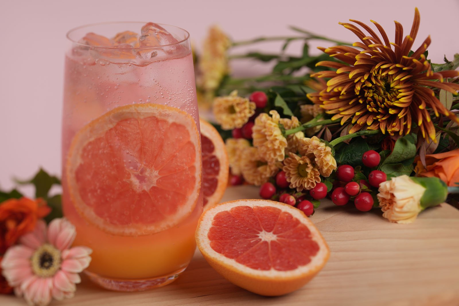 A refreshing grapefruit drink in a glass with ice, garnished with a grapefruit slice. Surrounding it are vibrant flowers, berries, and a halved grapefruit, all arranged on a wooden surface.