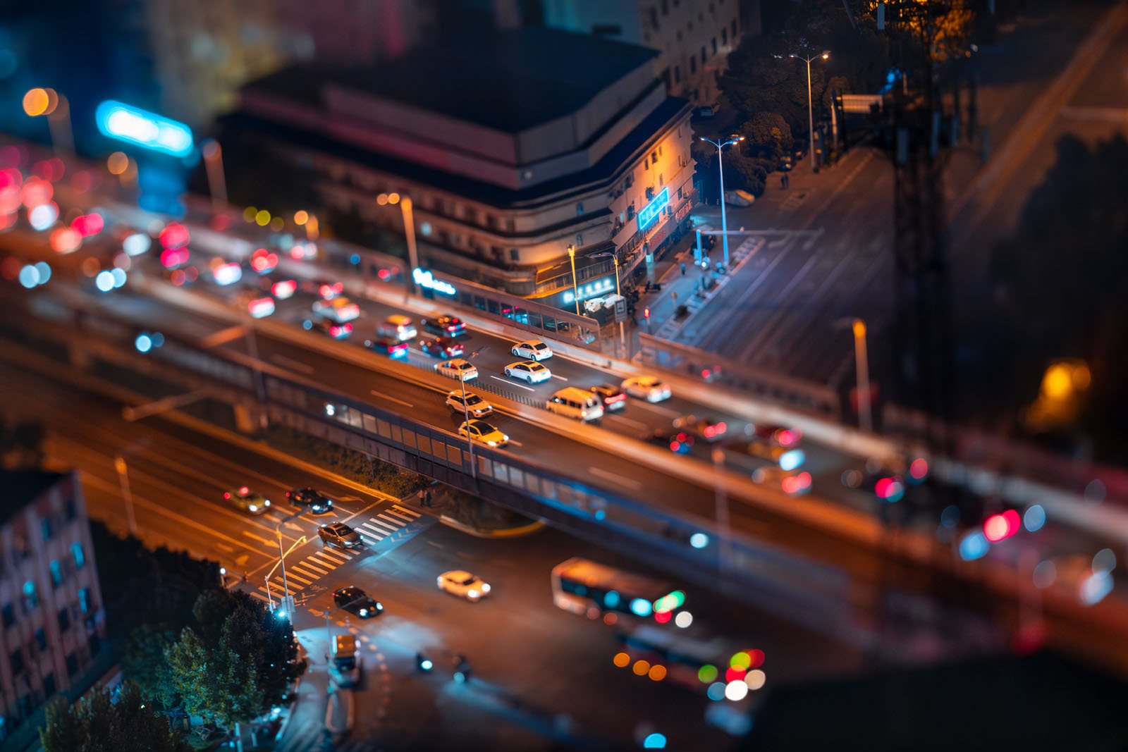 Aerial view of a busy city at night with bright, colorful lights. Traffic moves along streets and highways, creating a vibrant, bustling scene. Buildings line the roads, and blurred lights add to the dynamic, lively urban atmosphere.