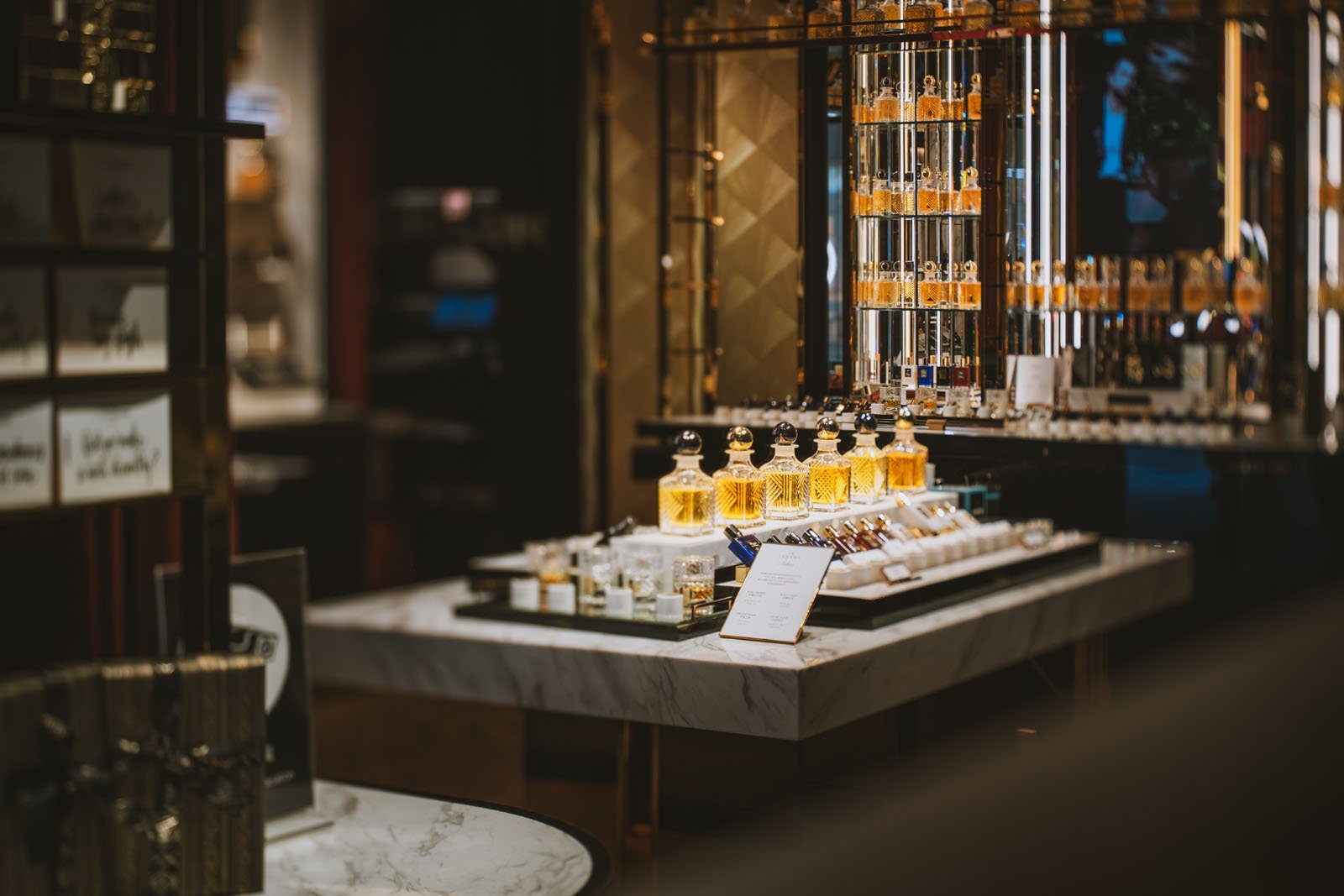 A luxurious perfume display in a store, featuring various bottles arranged on a marble table with warm lighting. Shelves with additional perfume bottles are visible in the background.