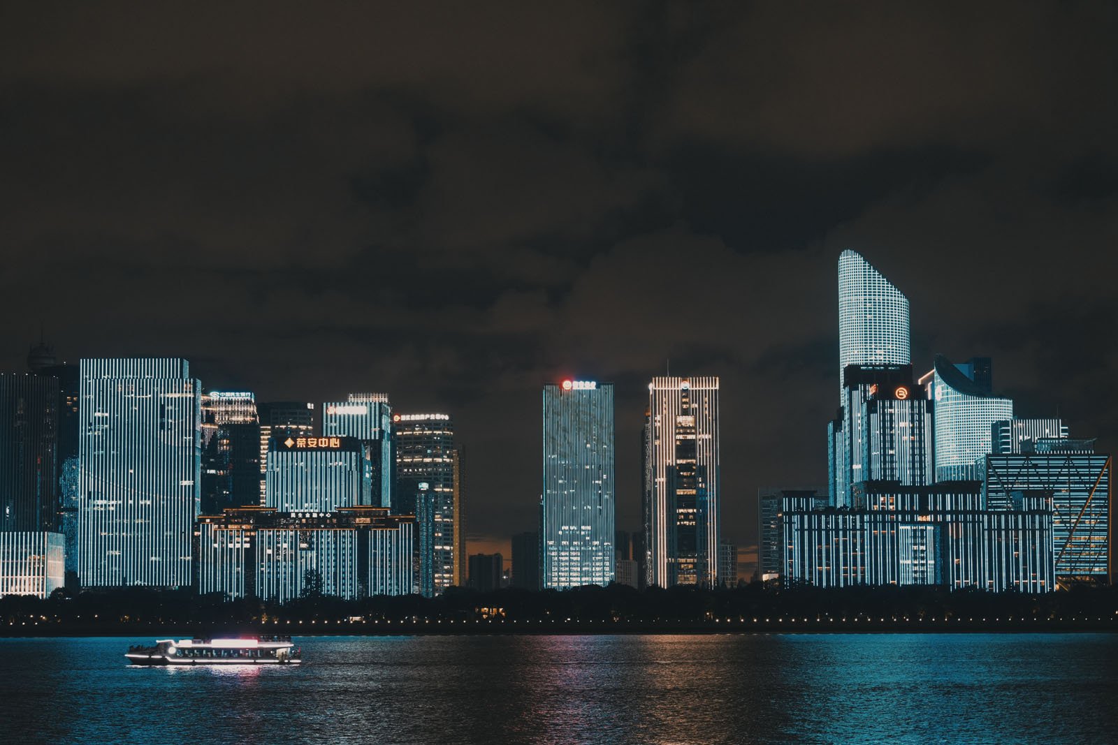 A nighttime cityscape featuring illuminated skyscrapers reflecting on a river. A brightly lit boat is cruising along the water, adding to the serene and vibrant urban scene.