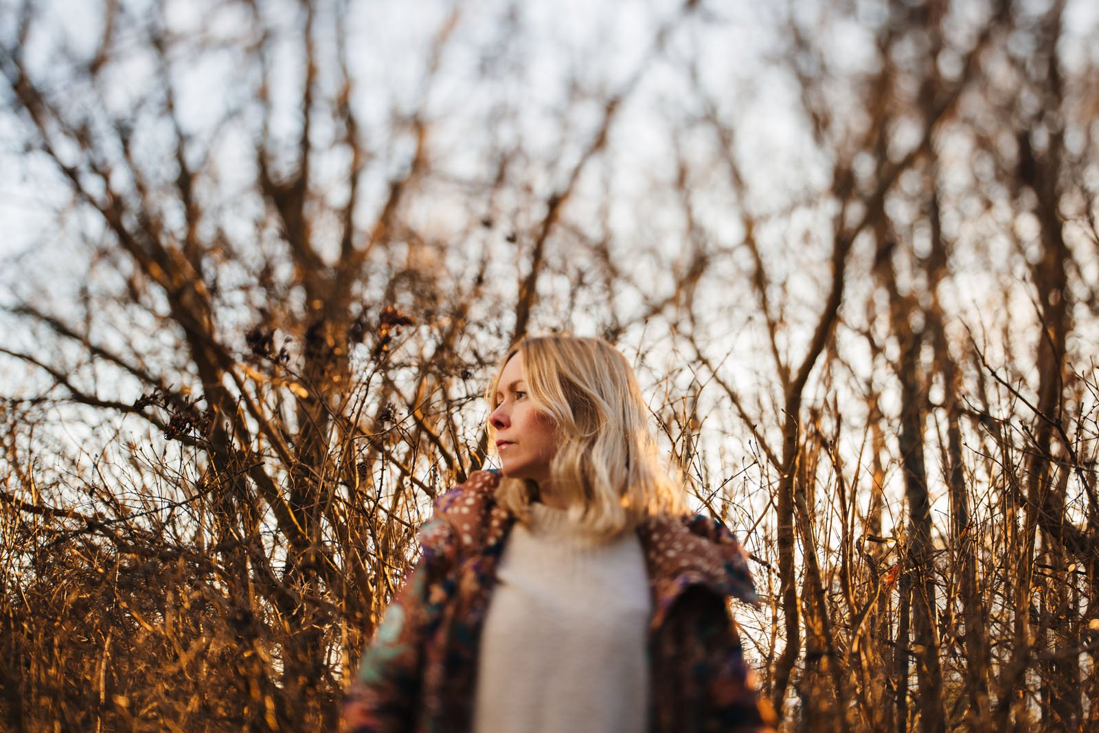 A woman with long blonde hair stands in a forest of leafless trees, wearing a patterned coat. The sun sets in the background, casting a warm glow on the scene. She gazes to the side, creating a thoughtful mood.