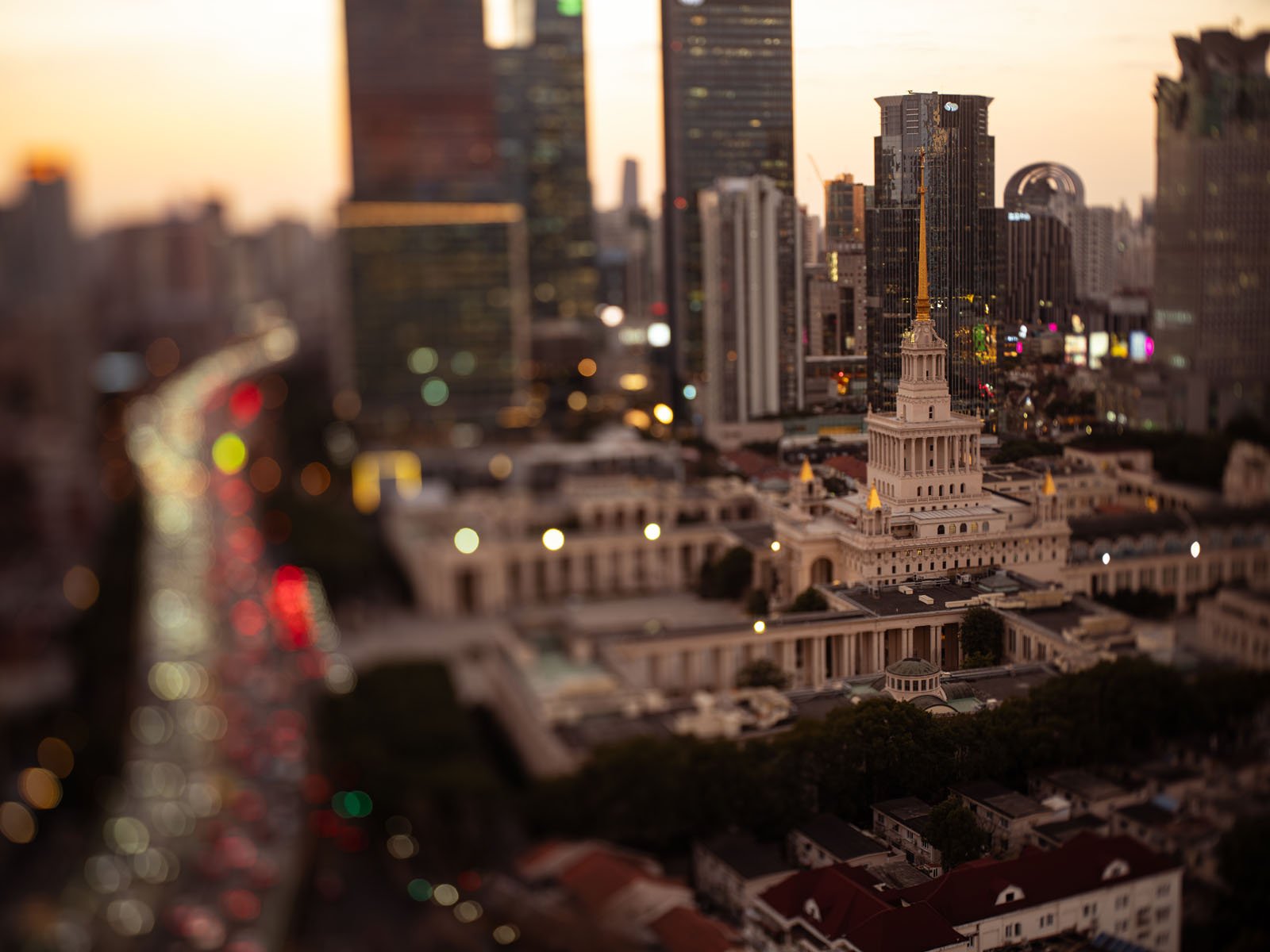 Tilt-shift image of a cityscape with a central white building with a golden spire, surrounded by tall skyscrapers. A busy road with blurred lights is visible to the left, creating a miniature effect.