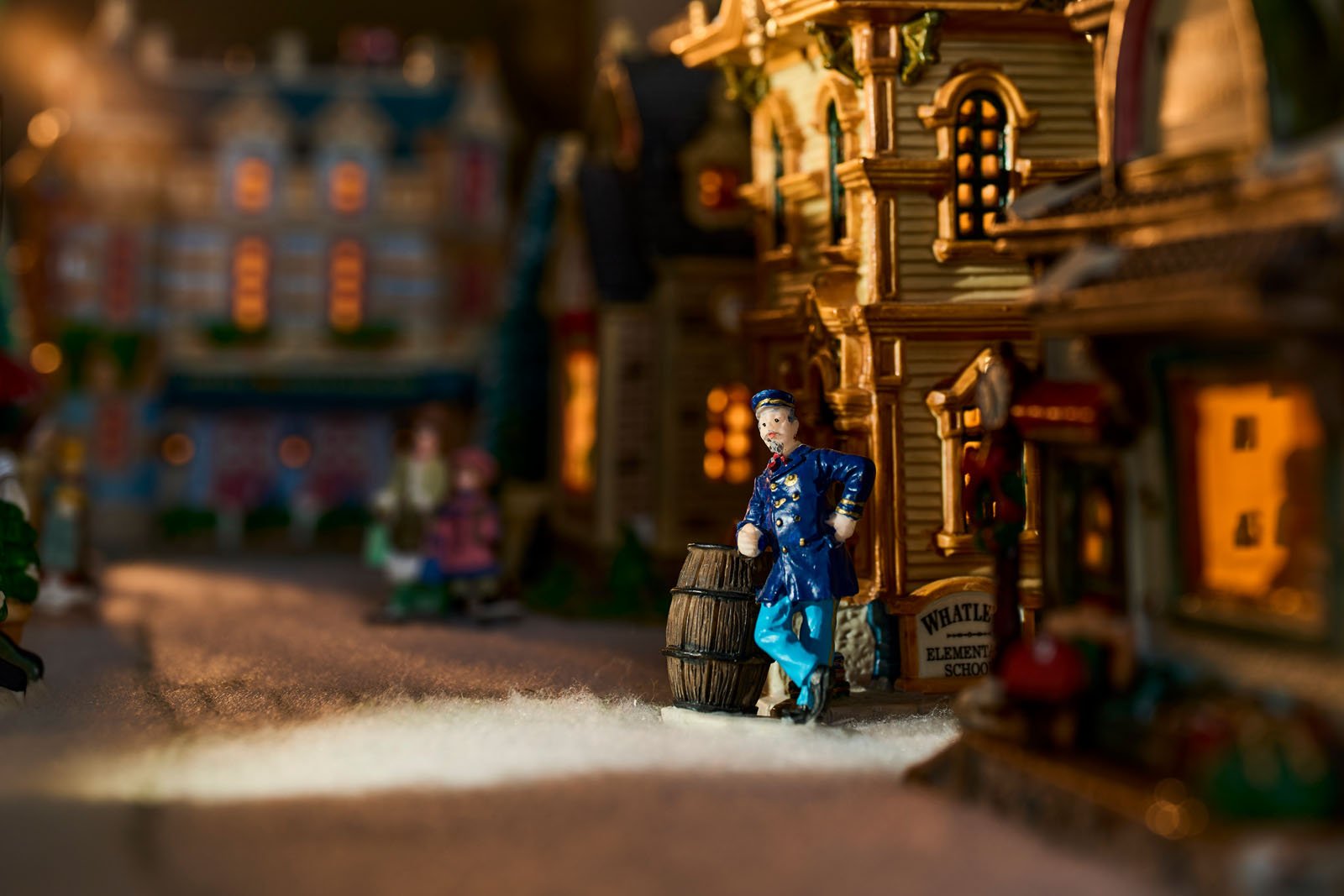 A miniature scene depicting a colonial-era village street, featuring a small figure of a woman standing by a barrel outside a shop called "Whalen's Biscuit Shop." Warm lighting highlights the richly detailed buildings and cobblestone road.