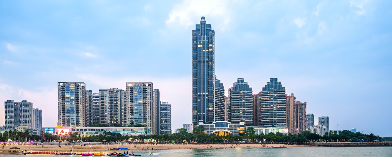 Panoramic view of a city skyline along a waterfront. The central towering skyscraper is flanked by several shorter buildings. People are visible on the beach, with a calm sea in the foreground and a cloudy sky.