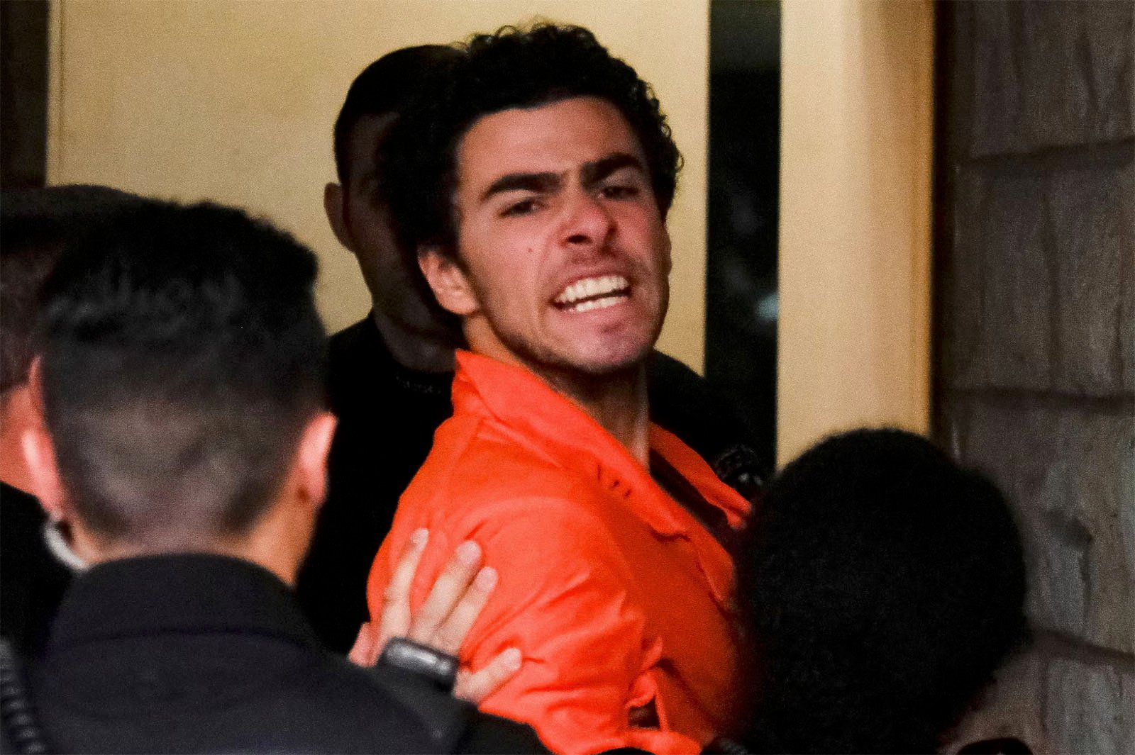 A person in an orange jumpsuit is being escorted by several people. They appear to be looking at the camera, with a tense expression. The setting seems to be indoors, near a doorway.