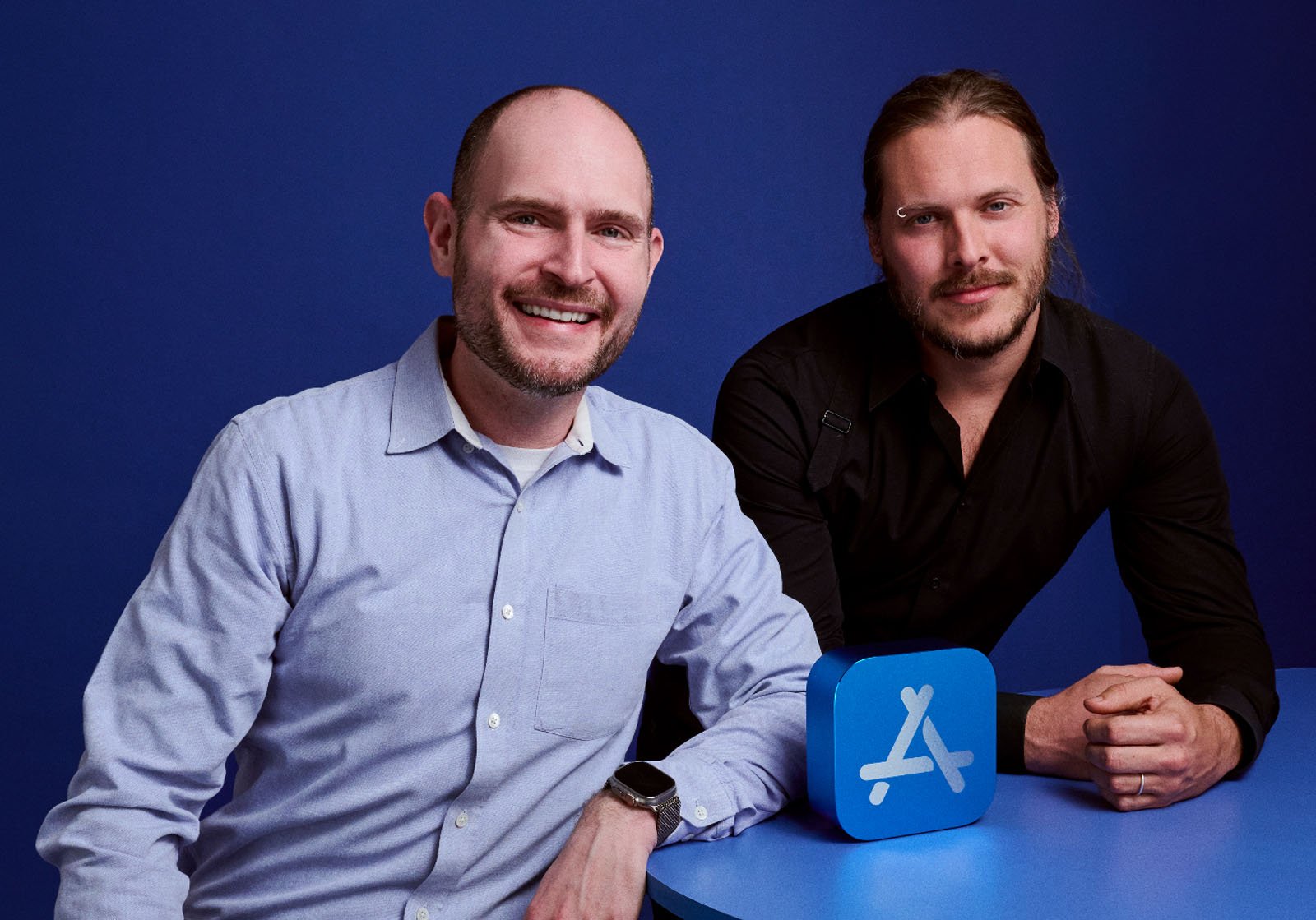 Two men smiling against a blue background. One is seated, wearing a light shirt and watch, the other leaning on a table, wearing a dark shirt. A blue cube with an app symbol is displayed on the table.