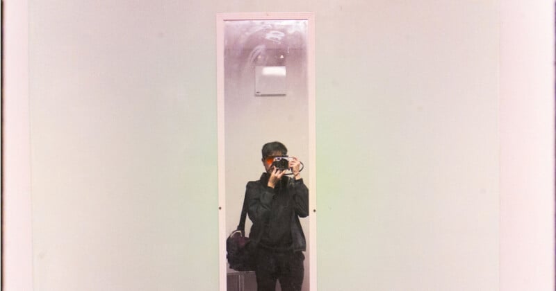 A person wearing dark clothing stands in front of a tall mirror, holding a camera that partially covers their face. The background is a plain, light-colored wall.