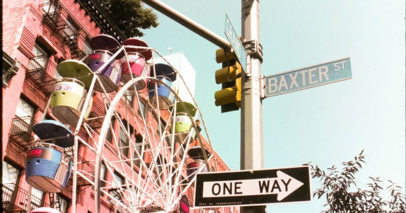 A colorful Ferris wheel beside a brick building is visible near a street sign reading "Baxter St." A one-way sign and a traffic light are also seen nearby, along with some tree branches.