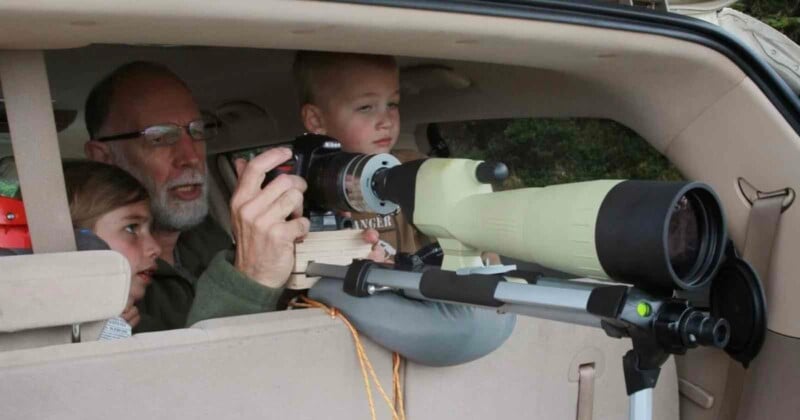 A man and two children sit in the back of a car with a camera and a large spotting scope mounted on the car window. They appear to be observing wildlife or birdwatching through the equipment.