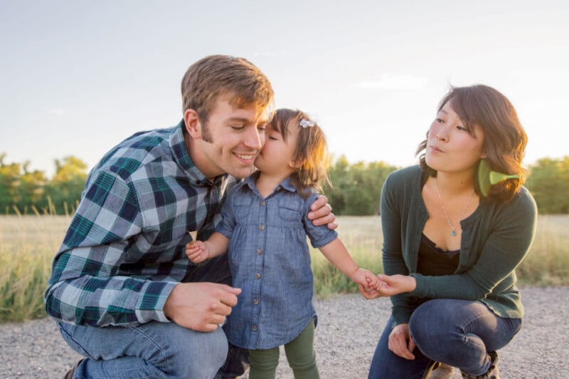 A young child kisses a smiling man on the cheek while holding the hand of a woman kneeling nearby. They are outdoors, with a grassy field and trees in the background, under a clear sky. The mood is joyful and affectionate.