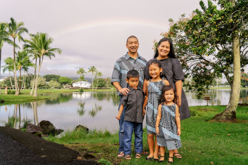 A family of five stands smiling near a tranquil lake with a faint rainbow in the background. All are dressed in matching blue patterned outfits. The scene includes palm trees, green grass, and a cloudy sky.