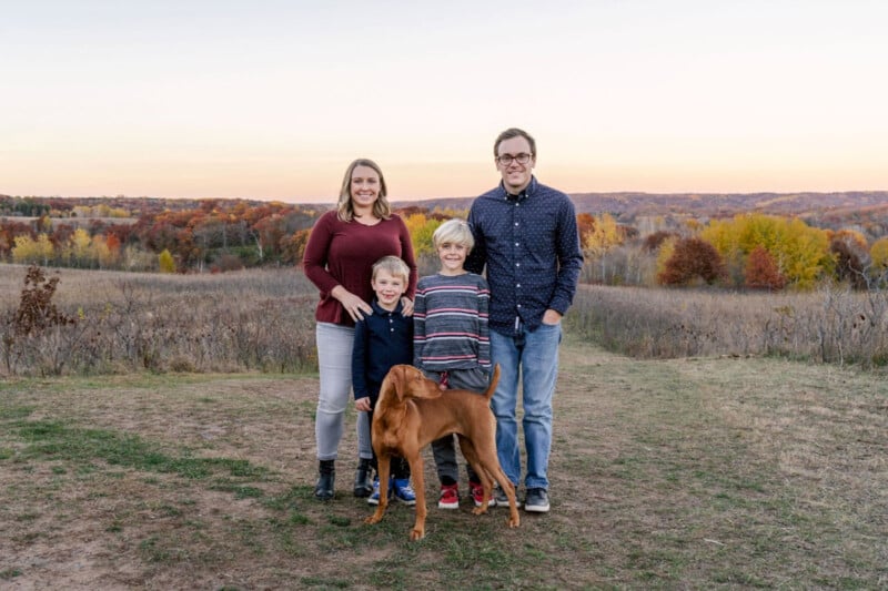 A family with a man, a woman, and two young boys stands outdoors in a field during autumn. A brown dog is with them. They are smiling, with trees displaying fall colors in the background under a clear sky.