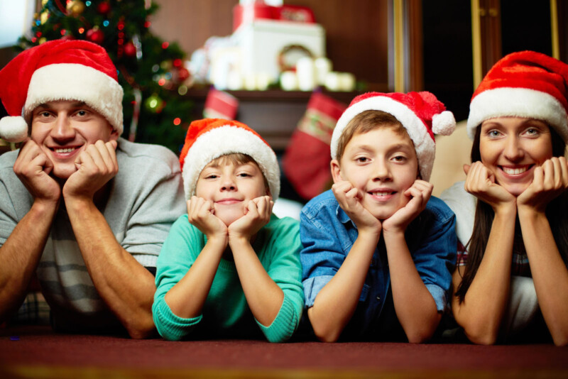 Four people wearing Santa hats are lying on the floor with their chins resting on their hands, smiling at the camera. A decorated Christmas tree is visible in the background. They appear to be a family enjoying a festive moment together.