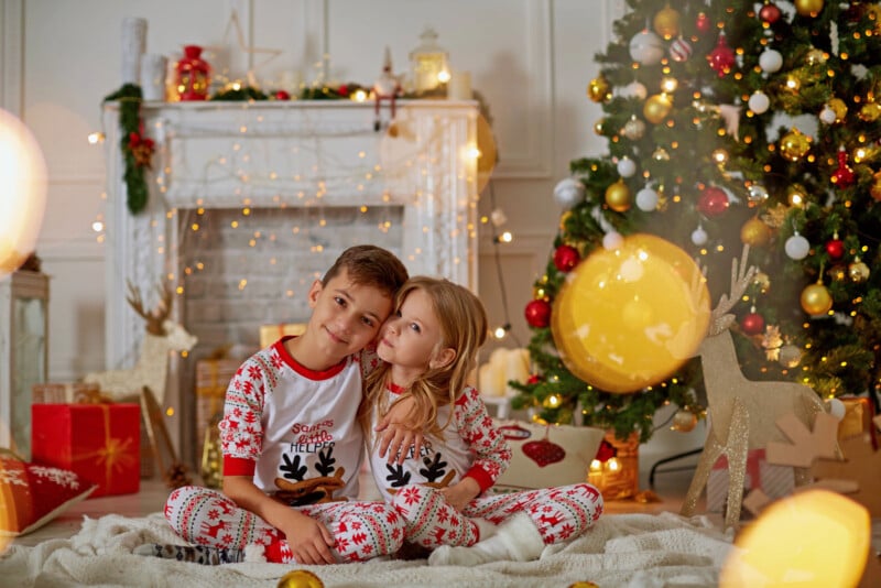 Two children in matching Christmas pajamas sit on a cozy rug in a festive living room. A decorated Christmas tree and gifts are behind them, along with a fireplace adorned with holiday decorations. Warm lights add a cheerful glow to the scene.