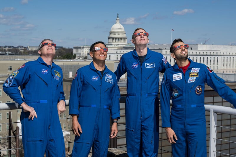 Four people in blue astronaut suits stand on a rooftop, wearing solar viewing glasses and looking up. The U.S. Capitol building is visible in the background under a partly cloudy sky.