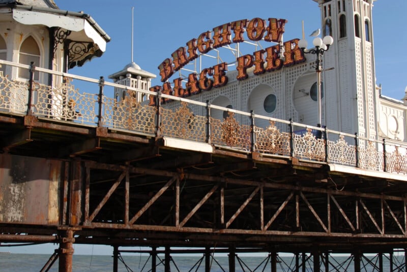 A view of the entrance to Brighton Palace Pier with its ornate sign and decorative railings, set against a clear blue sky. The pier’s structure is visible, highlighting metal beams and pillars above the sea.