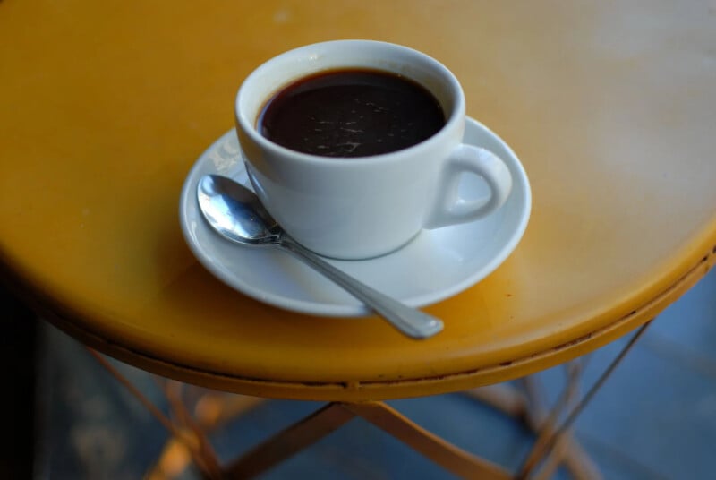 A white cup filled with black coffee sits on a matching saucer with a silver spoon beside it. The cup is placed on a round yellow table.