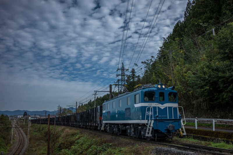 A blue and white freight train travels through a forested area under a cloudy sky. The train is on a track surrounded by green vegetation, with electric lines visible above. In the background, hills and trees can be seen.