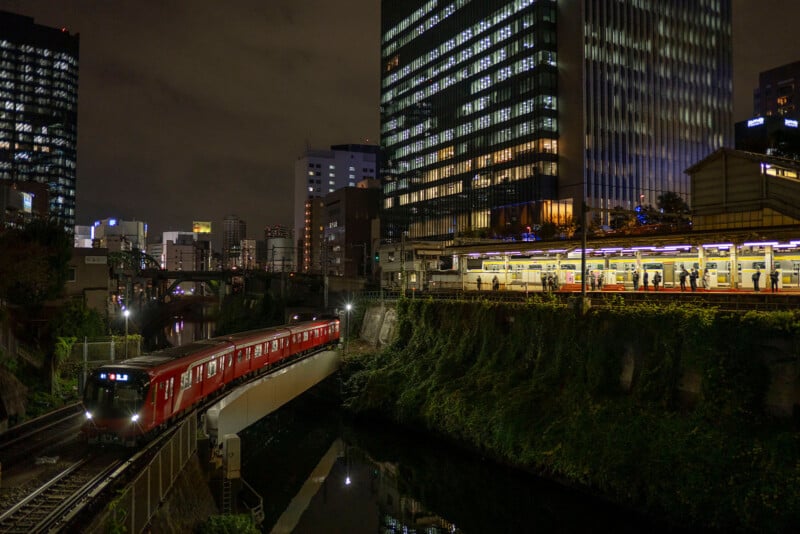 Nighttime urban scene featuring a red train crossing a bridge over a canal. To the right, a lit train station with passengers and a high-rise building with illuminated windows. The city skyline glows in the background under a cloudy night sky.