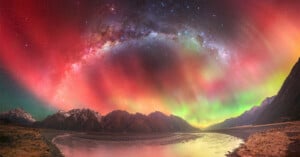 A breathtaking view of vibrant auroras in red, green, and purple hues lighting up the night sky over a mountainous landscape. A river flows through the scene, reflecting the colorful lights. The Milky Way is visible above.