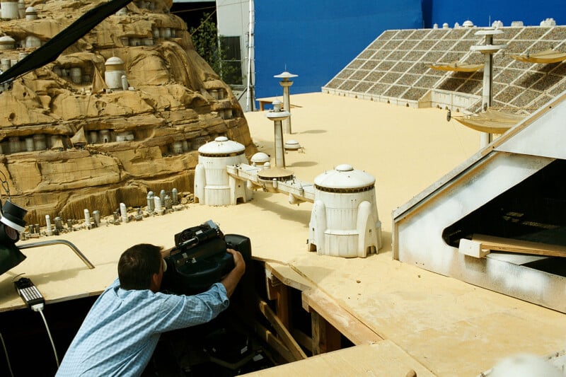 A person in a blue shirt operates a camera on a miniature desert city set, featuring beige terrain and futuristic white buildings, possibly for a film production.