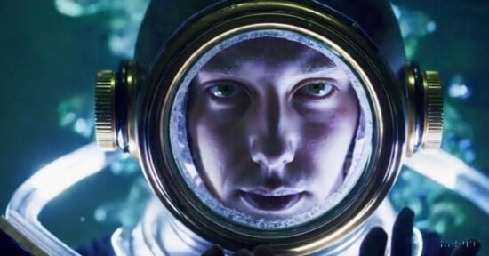 Close-up of an astronaut's face visible through a round helmet visor, surrounded by soft lighting that highlights their intense gaze. The background is blurred with a dark, ambient tone, adding a sense of mystery and exploration.