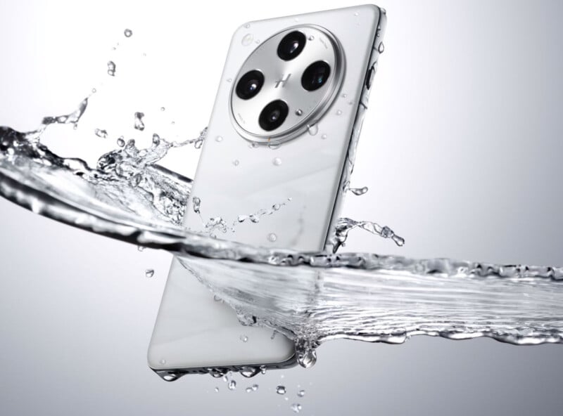 A sleek white smartphone is captured mid-air with water splashing around it, highlighting its water-resistant feature. The phone has a circular camera module with four lenses on the back. The background is a gradient of white and gray.