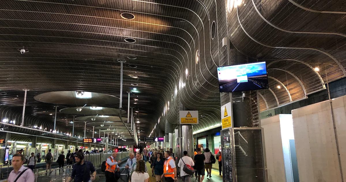 A bustling train​ station‌ with people walking in different directions. The ceiling is arched with ⁤a metallic design. Signs⁢ and digital screens are ⁤displayed‌ overhead,and workers in orange vests are visible among the travelers.